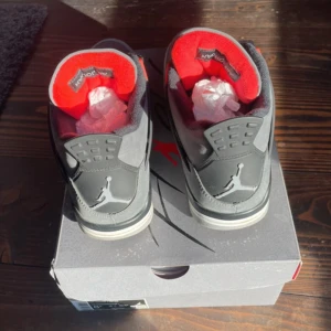 Nike Air Jordan 4 Retro Grå/Röd - Nike Air Jordan 4 Retro sneakers i grått med svarta och röda detaljer. Skorna har meshpaneler, snörning och Jumpman-logga på hälen och plösen. Snygg kontrast mellan den mörkgrå mockan och de röda accenterna. Platt sula med synlig Air-enhet för extra komfort.