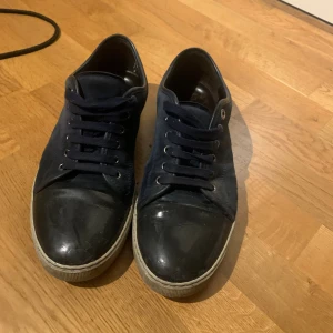 Mörkblå sneakers från Lanvin - Snygga mörkblå sneakers från Lanvin med ovandel i mocka och tåparti i svart skinn. Skorna har rund tå, platt vit sula och klassisk snörning. Perfekt för dig som gillar stilrena och exklusiva sneakers med en lyxig känsla.