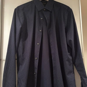Mörkblå prickig skjorta Banana Republic - Snygg mörkblå skjorta från Banana Republic med små vita prickar över hela tyget. Klassisk krage, knappar framtill och långärmad modell. Skjortan är tillverkad i bomull och har en clean, modern look som passar perfekt till jeans eller chinos.