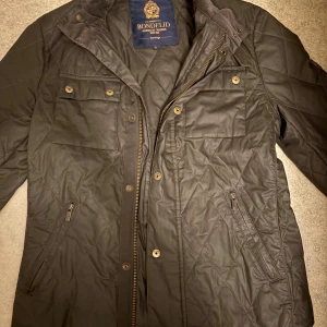 Svart quiltad jacka från Bondelid - Svart quiltad jacka från Bondelid med ståkrage och snyggt spänne vid halsen. Jackan har två bröstfickor med knappar, två snedställda fickor med dragkedja och stängs med både dragkedja och knappar. Perfekt för dig som gillar stilren och klassisk look. Mellan S och XS