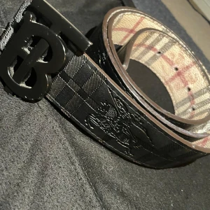Burberry vändbart bälte med B-spänne - Snyggt vändbart bälte från Burberry med klassiskt rutigt mönster på ena sidan i beige, rött och grått, och svart rutigt mönster med präglad ryttare på andra sidan. Stort svart B-spänne framtill. Tillverkat i skinn med exklusiv känsla och ikonisk design.