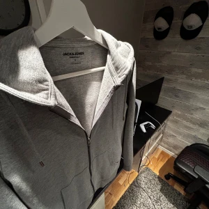 Grå zip hoodie - Säljer en stilren grå hoodie från Jack & Jones Essentials. Tröjan har dragkedja framtill, två fickor och justerbar huva med snören. Perfekt för chill dagar eller när du vill ha en enkel och clean look. Materialet är mjukt och skönt i bomullsmix.