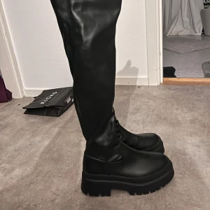 Svarta höga boots med grov sula - Säljer ett par svarta höga boots med chunky, räfflad sula och rund tå. Skorna är i syntetmaterial med dragkedja på insidan för enkel påtagning. Perfekta för dig som vill ha en edgy look och gillar att sticka ut med dina skor.