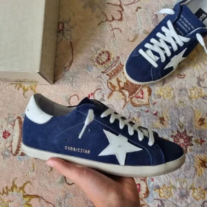 Blå Golden Goose sneakers med stjärna - Snygga sneakers från Golden Goose i mörkblå mocka med vit stjärndetalj på sidan och vit häl. Skorna har vita snören och platt sula. Kommer med originalkartong och dustbag. Perfekta för dig som gillar streetstyle och vill sticka ut.