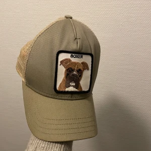 Beige keps Goorin Bros Boxer mesh - Snygg beige keps från Goorin Bros med mesh-bakstycke och broderad patch framtill med en boxerhund och texten 'BOXER'. Kepsen har böjd skärm och justerbar passform. Perfekt för dig som gillar streetstyle och djurmotiv.