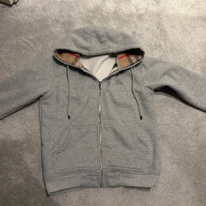Grå Burberry hoodie med rutig huva - Säljer en grå hoodie från Burberry med dragkedja och klassiskt rutigt foder i huvan. Tröjan har snörning i huvan, två fickor fram och ribbade muddar. Tillverkad i mjuk bomullsmix, perfekt för chill dagar. För endast 600kr i storlek S hör av er vid frågor. 10/10 skick.