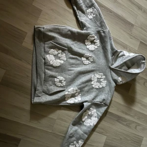 Grå hoodie från Denim Tears med blommor - Grå hoodie från Denim Tears med vita blommiga applikationer över hela tröjan. Tröjan har huva, magficka och är tillverkad i mjuk bomullsblandning. Perfekt för dig som gillar streetwear med unik design. Lite baggy passar även M för de e lite baggy perfekt nu för vinter. Den är väldigt varmt. Pris kan diskuteras vid snabb affär. 