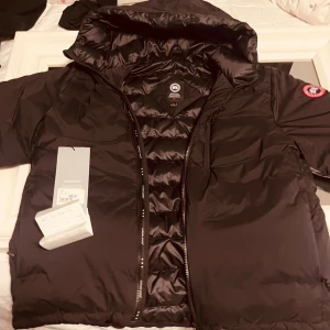 Svart dunjacka från Canada Goose - Säljer en svart dunjacka från Canada Goose med huva och dragkedja. Jackan har klassisk logga på ärmen, flera fickor med dragkedja och är tillverkad i polyester med dunfyllning. Perfekt för kalla dagar och riktigt snygg passform. 9,5/10 skick