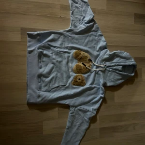 Ljusblå Palm Angels hoodie med björn - Ljusblå hoodie från Palm Angels med luva och vit snörning. På bröstet finns ett broderat motiv av en nallebjörn med avtagbart huvud. Tröjan har en stor magficka och är i mjuk bomull. Perfekt för dig som gillar streetwear och unika detaljer. Lite baggy passar S/M storlek står inte men de stog när ja köpte den. Köpte i fel storlek och det är därför jag säljer den har aldrig använt den. 