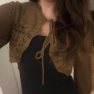 Stickad brun väst med snörning - Säljer en kort, stickad väst i brun nyans med snyggt virkat mönster och långa ärmar. Västen knyts framtill med snörning och har en croppad passform som ger en trendig look. 