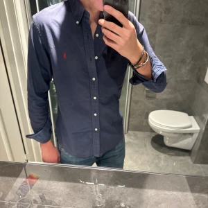 Mörkblå skjorta från Ralph Lauren - Snygg mörkblå skjorta från Ralph Lauren med klassisk button-down krage och vita knappar framtill. Skjortan är I riktigt bra skick och har inga defekter. Jag är 188cm och bör storlek M på bilden. Hör av er vid funderingar:)