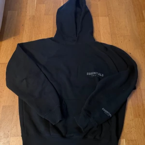 Svart Essentials Fear of God hoodie - Svart hoodie från Essentials Fear of God med tryckt logga på bröstet och ärmen. Tröjan har huva, känguruficka och långa ärmar. Materialet är mjuk bomull och passformen är loose, perfekt för en chill och avslappnad stil.