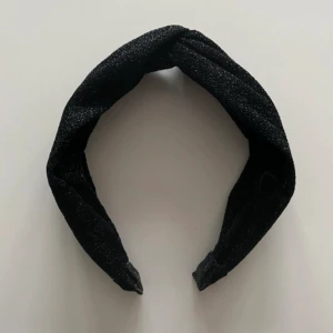 Svart glittrigt diadem med twist - Snyggt svart diadem med glittrigt tyg och twistad detalj på ovansidan. Perfekt accessoar för att lyfta din look med lite extra shimmer. Fint skick!