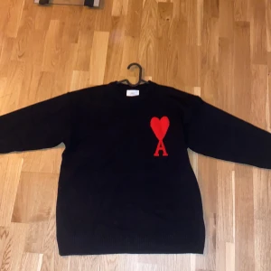 Svart stickad tröja från Ami Paris - Svart stickad tröja från Ami Alexandre Mattiussi med lång ärm och rund hals. På bröstet finns ett rött hjärta och bokstaven A som broderad detalj. Perfekt för dig som gillar stilrena plagg med en twist.