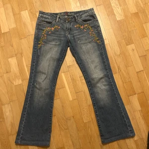 Blå bootcut Lågmidjade jeans - Snygga blå bootcut jeans med broderade blommor i rosa och gult vid fickorna. Jeansen har klassisk femficksdesign, slitningar och broderad logga i linningen. Perfekt för dig som gillar detaljer och en avslappnad stil. Midjemåttet är 41cm