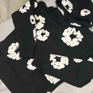 Svart hoodie från Denim Tears med tryck - Svart hoodie från Denim Tears med stora vita blommönster tryckta över hela tröjan. Tröjan har huva och långa ärmar, samt en klassisk loose passform. Materialet är mjuk bomull som känns skön mot huden. Perfekt för dig som gillar streetwear och unik design.