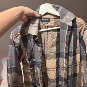 Rutig skjorta från Barbour - Snygg rutig skjorta från Barbour med klassiskt tartanmönster i beige, grått, svart och röda detaljer. Skjortan har lång ärm, krage och knappar framtill. Perfekt för dig som gillar tidlös stil och vill ha något som sticker ut lite extra.