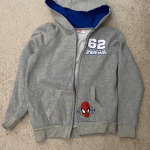Grå Spiderman hoodie med dragkedja - Säljer en grå hoodie med blått foder i huvan och coolt Spiderman-tryck på bröstet och fickan. Tröjan har dragkedja framtill, två fickor och ribbade muddar. Perfekt för dig som gillar superhjältar och vill ha en bekväm och snygg stil.