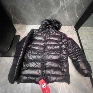 Canada goose jacka - Säljer en svart dunjacka från Canada Goose med klassisk logga på ärmen. Jackan har en puffad design, huva och två dragkedjeförsedda fickor framtill. Perfekt för kalla dagar och har en riktigt snygg streetstil. Äktenhet kan ej garanteras men antagligen Äkta