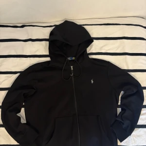 Svart hoodie från Polo Ralph Lauren - Svart hoodie från Polo Ralph Lauren med klassisk broderad logga på bröstet. Tröjan har huva med snörning, dragkedja framtill och två fickor. Materialet är mjuk bomull och passformen är normal. Perfekt för chill dagar och enkel att matcha med jeans eller joggers. Hör av dig om du har några frågor.