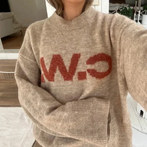 Beige stickad tröja med röd text - Beige stickad tröja med oversized passform och breda ribbade muddar. Framsidan har en stor röd text i grafisk stil. Tröjan har rund halsringning och långa ärmar, perfekt för lager-på-lager under kyliga dagar.