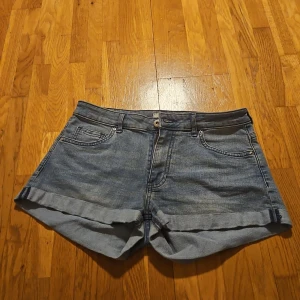 Blå jeansshorts från H&M, strl 36 - Snygga blå jeansshorts från H&M i klassisk femficksmodell med uppvikta ben. De har normal passform och midwaist, perfekt för sommaren. Materialet är slitstark bomull och shortsen har knapp och dragkedja framtill.