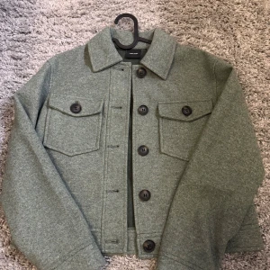 Grön jacka från Vero Moda - Grön jacka från Vero Moda med stora svarta knappar och två bröstfickor med lock. Tillverkad i ett mjukt ullblandat material. Perfekt lager-på-lager-plagg för kyligare dagar. Använd få gånger, som ny. 