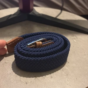 Mörkblå flätat bälte med läderdetaljer - Snyggt mörkblått flätat bälte med bruna läderdetaljer och silverspänne. Bältet har en stilren design och passar perfekt till jeans eller chinos. Den flätade strukturen ger en modern och avslappnad känsla.