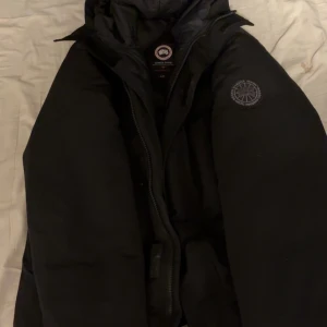 Svart dunjacka från Canada Goose L - Säljer en svart dunjacka från Canada Goose i storlek L. Jackan har klassisk pufferdesign, huva och dragkedja framtill. Ikoniska Canada Goose-loggor syns på ärmen och insidan. Perfekt för kalla vinterdagar och riktigt snygg streetstil.