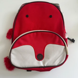 Röd ryggsäck med rävmotiv och fluffiga detaljer - Supergullig röd ryggsäck med rävansikte framtill, vita och bruna detaljer samt fluffiga röda tofsar på dragkedjorna. Väskan har justerbara axelband, sidoficka med dragsko och ett stort fack framtill. Perfekt för dig som vill sticka ut! 