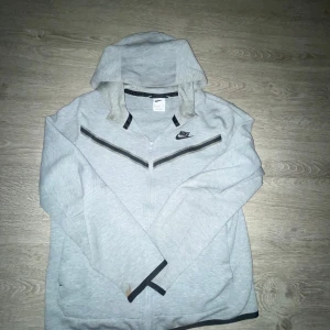 Grå Nike hoodie med dragkedja XL - Snygg ljusgrå hoodie från Nike i storlek XL med svart Nike-logga på bröstet och svarta detaljer längs dragkedjan och muddar. Tröjan har huva och hel dragkedja framtill, perfekt för en sportig och avslappnad stil.