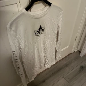 Vit långärmad Adidas tröja - Säljer en vit långärmad tröja från Adidas med svart logga på bröstet och tryck längs ena ärmen. Tröjan har en klassisk rund halsringning och är gjord i mjuk bomull, perfekt för en sportig och avslappnad stil.