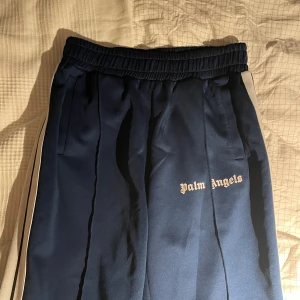 Blå Palm Angels byxor - Säljer ett par mörkblå byxor från Palm Angels med vita sidoränder och logga på låret. Byxorna har elastisk midja och raka ben. Har haft byxorna i 3 år och är använda en del men mest som mjukisbyxor hemma bara. Inga fläckar inga defekter.