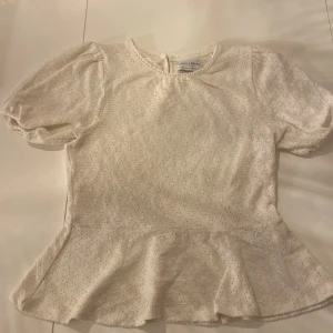 Vit blus med volang från Lindex - Supersöt vit blus från Lindex i storlek S. Blusen har korta puffärmar, volang nertill och ett diskret hålmönster i tyget. Rund halsringning med liten knapp i nacken. 