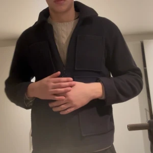 Svart ulljacka från mfpen - Svart jacka i ull med fyra stora fickor framtill och dragkedja. Klassisk krage och rak passform, perfekt för dig som gillar stilren och funktionell design. Jackan är långärmad och har en clean look utan synliga loggor.