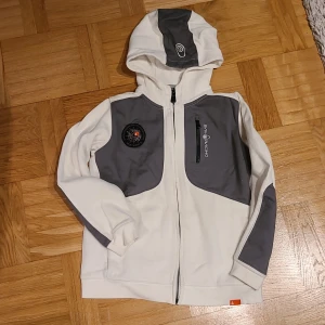 Vit och grå hoodiejacka Sail Racing - Vit och grå hoodiejacka från Sail Racing med dragkedja framtill, huva och gråa detaljer på axlar, bröst, armbågar och huva. Jackan har en bröstficka med dragkedja och Sail Racing-logga samt en patch på bröstet. Perfekt för dig som gillar sportig stil.