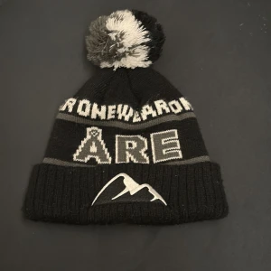 Svart och grå Åre-mössa med tofs - Stickad svart och grå mössa från onewear med stor tofs i svart, grått och vitt. Framtill står det 'ÅRE' i stora bokstäver och ett broderat bergmotiv. Perfekt för kalla dagar och vintersäsongen.