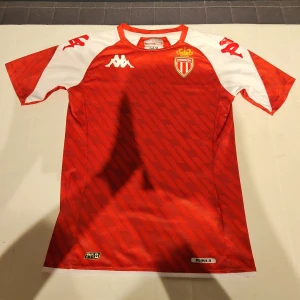 AS Monaco röd/vit fotbollströja Kappa - AS Monaco matchtröja från Kappa i rött och vitt med diagonalt mönster. Kortärmad modell med klubbmärke på bröstet och Kappa-loggor på axlarna. Tillverkad i lätt och ventilerande polyester, perfekt för träning eller match. Köpte den när jag var på semester i Monaco och nu så passar den inte mig längre så nu ska jag sälja den.