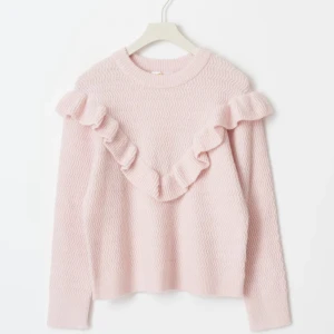 Ljusrosa stickad tröja med volang - Superfin ljusrosa stickad tröja med volangdetaljer framtill och på axlarna. Tröjan har ett subtilt vågmönster i materialet och rund hals. Perfekt för dig som gillar söta och trendiga detaljer.