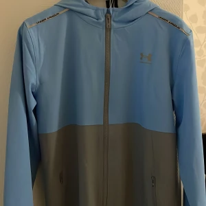 Blå och grå vindjacka från Under Armour - Snygg tvåfärgad vindjacka från Under Armour i ljusblått och grått. Jackan har huva, hel dragkedja framtill och diskret logga på bröstet. Perfekt för träning eller när du vill ha en sportig look. Långa ärmar och praktiska fickor med dragkedja.