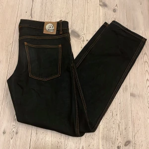 Cheap Monday Jeans  - Svarta jeans från ”Cheap Monday” med orange kontrastsömmar och unik dödskalle-patch bak i midjan. Innerfoder med tryckta dödskallar i ljusgrönt. Klassisk femficksmodell med rak passform och dragkedja. Perfekt för dig som vill sticka ut med coola detaljer.