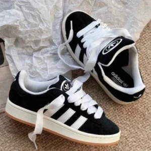 Adidas Campus svartvita sneakers - Säljer ett par Adidas Campus sneakers i svart mocka med vita detaljer och klassiska tre ränder på sidan. Skorna har vit sula med gummidetalj och breda vita skosnören. Perfekta för dig som gillar retrostil och streetwear. De är super fina och kan gå ned i pris vid snabb affär 