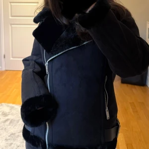 Svart mocka jacka med päls - Snygg svart pälsjacka från Pull&Bear krage och mjuk fuskpäls på insidan och vid ärmslut. Jackan har silvriga dragkedjor, spänndetaljer vid kragen och midjan samt en cool, lite oversized passform. Aldrig använd! Prislapp är kvar. 