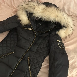 Svart dunjacka Rockandblue med päls - Snygg svart dunjacka från Rockandblue med guldiga dragkedjor och quiltade detaljer. Jackan har en stor huva med fluffig ljus päls, flera fickor med dragkedja och ett coolt Rockandblue-emblem på ärmen. Perfekt för kalla dagar och riktigt stilren look.