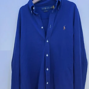 Blå skjorta från Ralph Lauren M - Snygg blå skjorta från Ralph Lauren med klassisk button down-krage och broderad polospelare på bröstet. Skjortan är i bomull och har långa ärmar samt vita knappar framtill. Perfekt för dig som gillar ”grisch” stilen.
