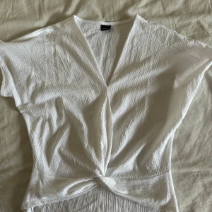 Vit omlottopp från Gina Tricot - Säljer en vit omlottopp från Gina Tricot i storlek XS. Toppen har korta ärmar, v-ringning och en snygg twistad detalj framtill. Materialet är lätt strukturerat och känns luftigt, perfekt för varmare dagar.