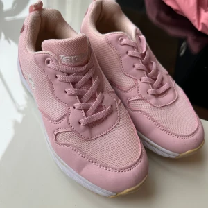 Rosa sneakers från Kappa - Snygga rosa sneakers från Kappa med vit logga på sidan. Skorna har snörning, platt sula och är gjorda i en mix av syntet och textil. Perfekta för dig som vill ha en sportig och trendig look med en touch av färg.