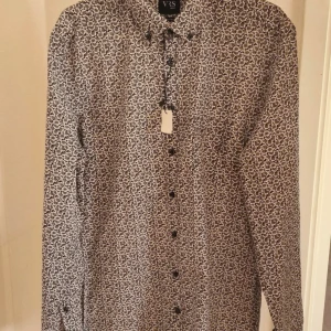 Blommig skjorta från VRS - Mönstrad skjorta från VRS med små blommor i svart och vitt. Skjortan har lång ärm, knappar framtill och klassisk krage. Perfekt för dig som gillar detaljerade mönster och stilren design.