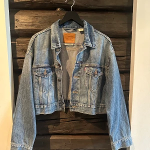Levi's jeansjacka i klassisk blå denim - Säljer en klassisk blå jeansjacka från Levi's Premium i modell med raka linjer och croppad passform. Jackan har två bröstfickor med knappar, knäppning framtill och typisk denimlook. Perfekt för dig som gillar tidlös streetstyle. Använd ett fåtal gånger. 