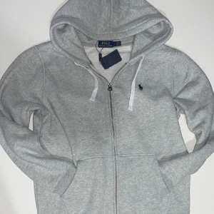 Grå hoodie från Polo Ralph Lauren - Snygg grå hoodie från Polo Ralph Lauren med dragkedja och huva. Tröjan har vita snören, broderad logga på bröstet och mjukt material. Perfekt för en chill och stilren look. Storlek L men passar även M